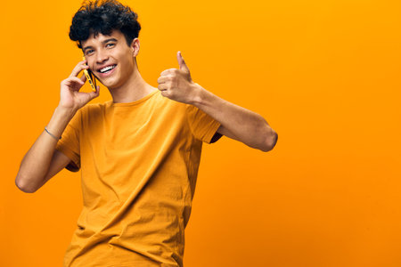 Young man smiling while talking on the phone, giving a thumbs up gesture against a bright yellow background, conveying positivity and happiness in a cheerful momentの写真素材