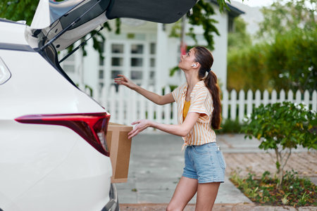 Woman unloading a box from the trunk of a car, outdoor setting with greenery and a white picket fence in background, sunny day, casual clothingの写真素材