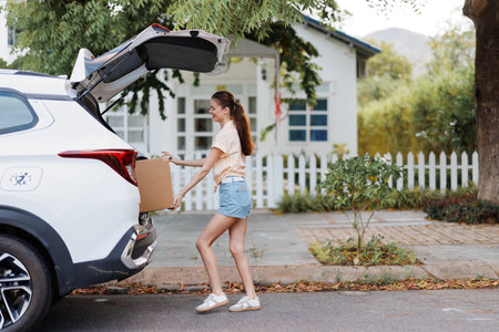 woman unloading a cardboard box from the trunk of a car in a residential area with greenery and a white picket fence in the background, bright and sunny environment, street sceneの写真素材