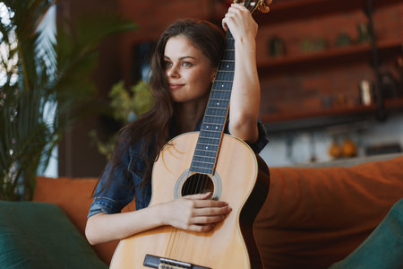 Young woman sitting on a couch holding an acoustic guitar in front of her face and looking at the cameraの写真素材