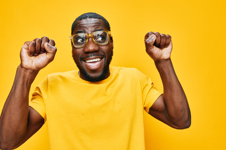 Happy joyful young man celebrating success with raised fists, wearing stylish glasses and a yellow t shirt against a bright yellow backgroundの写真素材