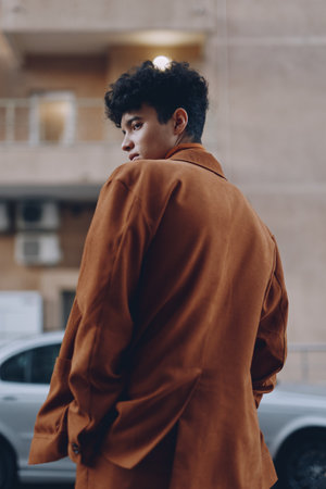 Young man in a brown coat looking back with a thoughtful expression against an urban backdropの写真素材