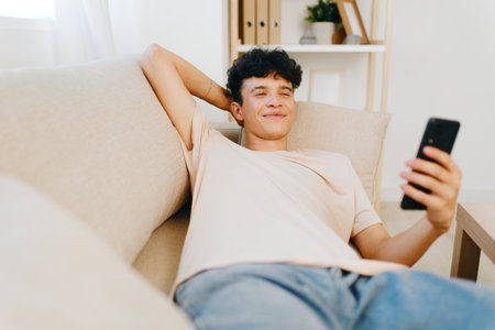 Relaxed young man using smartphone on a cozy couch, casual attire, indoor setting with light colors for a calm atmosphere Smiling face conveys leisure and comfortの写真素材