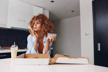 Woman enjoying a casual meal with a box of pizza and a bag of chips at a tableの写真素材
