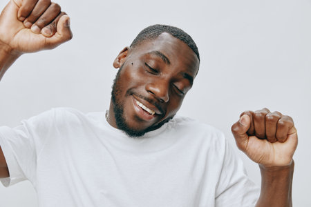 Happy African male celebrating life, wearing a casual white t shirt, smiling with arms raised, conveying joy and excitement in a simple light backgroundの写真素材