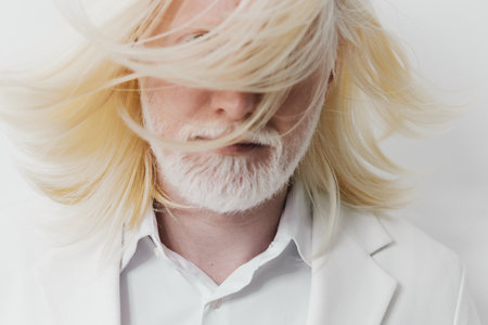 Creative portrait of an androgynous man with long blonde hair and beard, wearing a white suit against a minimalistic white background, conveying a sense of modern elegance and styleの写真素材