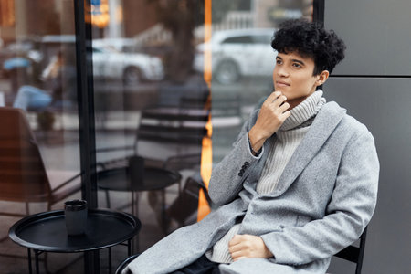 Fashionable young man in a gray coat sitting by a window holding a cup of coffeeの写真素材