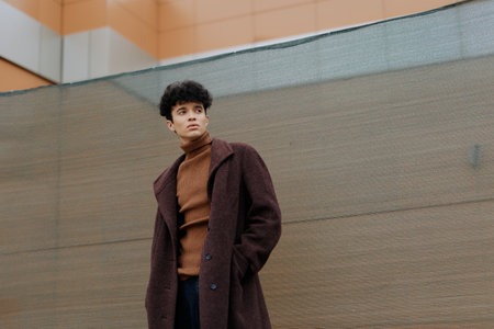 Fashionable young man in brown coat and turtleneck sweater standing with hands in pockets in front of wallの写真素材