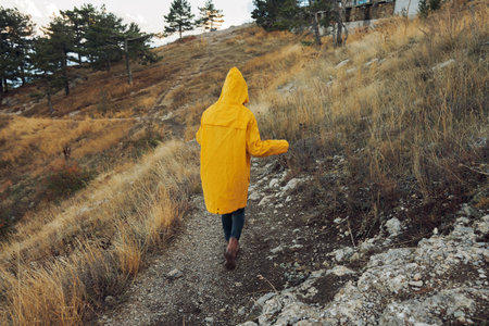 A woman wearing a yellow raincoat walking down a trail in the mountainside during a scenic mountain hikeの写真素材