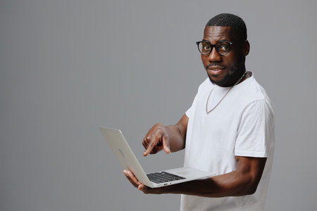 African man smiling while pointing at laptop, wearing white T shirt, casual attire, gray background, expressing confidence and approachability in a modern settingの写真素材
