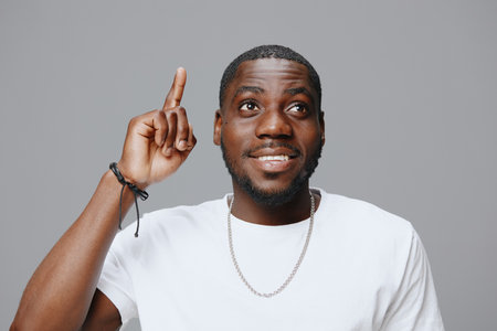 smiling young man with raised finger, casual white t shirt, showing idea or inspiration, neutral gray background, expressing happiness and confidence, perfect for creative concepts related to innovatの写真素材