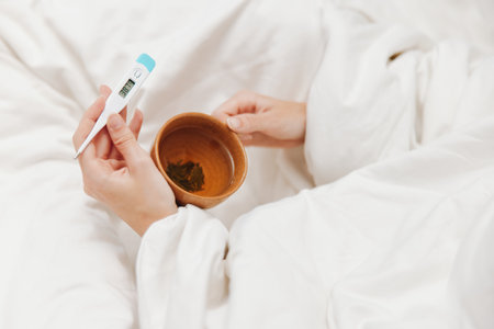 Ill person in bed holding a thermometer and a cup of teaの写真素材