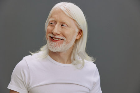 Happy elderly man with long white hair and beard smiling against a neutral gray background,...