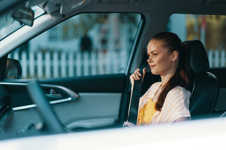 Young woman sitting in a car, smiling, wearing a yellow dress, softly lit interior, casual look, city background out of focus for lifestyle imageryの写真素材