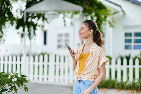 Young woman using smartphone outdoors, wearing casual attire, standing against a bright, sunny...