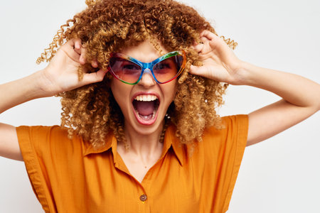 Happy woman with curly hair expressing joy, wearing colorful sunglasses, against a plain white background, showcasing vibrant fashion and emotionの写真素材