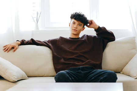 Young man sitting on a cozy sofa in a bright room, relaxed posture with a thoughtful expression, soft natural light filtering through the windowsの写真素材