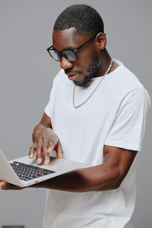 Black man using laptop, casual clothing, modern technology, neutral background, focused expression,...