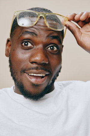 Cheerful young man adjusting eyewear, expressing surprise and joy, wearing a casual white t shirt, with a neutral light colored background that complements his emotionsの写真素材
