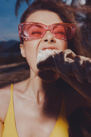 Young woman enjoying an ice cream cone in stylish pink glasses, vibrant yellow swimsuit, against a tropical background with mountains and palm treesの写真素材