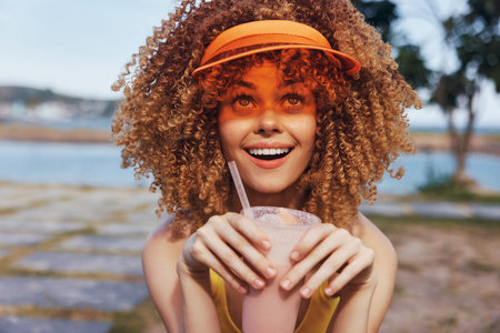 Cheerful young woman with curly hair holding a smoothie, wearing a bright visor, smiling against a natural outdoor backgroundの写真素材