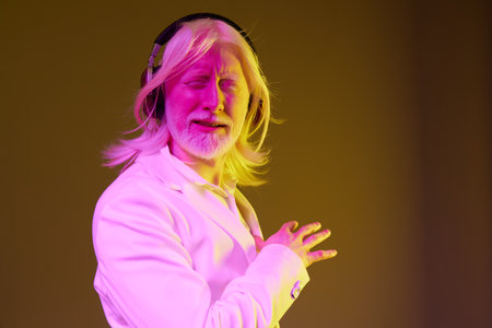 Portrait of a stylish older man with long white hair and headphones against a vibrant yellow...