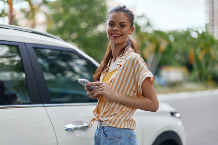 Young woman in a casual outfit holding a smartphone while standing next to a white car, smiling...