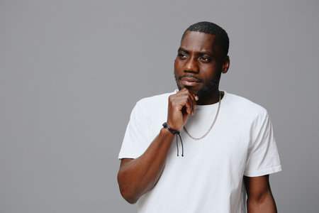 Thoughtful African man in casual white t shirt contemplating with a serious expression against a neutral gray background The model exudes confidence and introspectionの写真素材