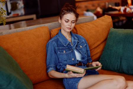 Woman reading book on orange couch in denim jacket cozy home interior relaxing leisure activity peaceful momentの写真素材