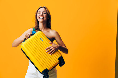 Happy woman with a yellow suitcase standing in front of vibrant orange wall with smile on her faceの写真素材