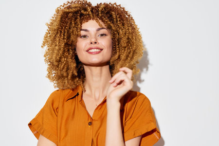 Smiling woman with curly hair in a stylish orange shirt against a bright white background, exuding...