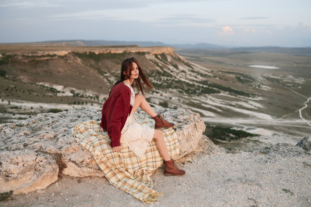 Woman enjoying peaceful moment on cliff edge overlooking scenic landscape with blanket and scarf, travel and relaxation conceptの写真素材