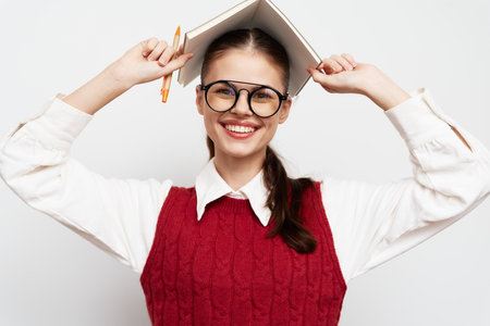 Happy young woman holding a book above her head, wearing glasses and a red sweater against a light gray background, portraying joy and enthusiasm for learning, lively expressionの写真素材