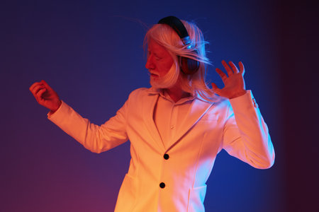 Creative elderly man with headphones dancing against a vibrant gradient background, showcasing joy...