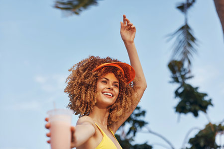 Happy woman with curly hair enjoying a drink in a tropical setting, wearing a bright yellow swimsuit and orange visorの写真素材