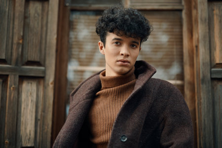 Fashionable young man with curly hair wearing a brown coat and turtleneck sweater, standing in front of an old wooden door, looking to the sideの写真素材