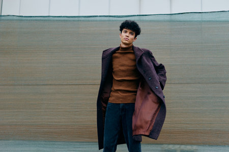 Fashionable young man in brown sweater and coat, blue jeans, black boots, standing in front of a wallの写真素材