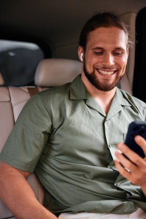 Man sitting in a car, smiling while using a smartphone, with earbuds in, dressed in a green shirt, showcasing a relaxed atmosphereの写真素材