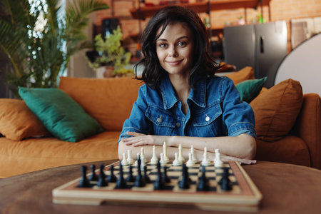 Woman sitting on couch in front of chess board with hands on hips in thoughtful poseの写真素材
