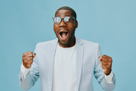 Happy African man celebrating success with enthusiasm in a stylish light suit on a blue background, expressing joy and excitement with a big smile and raised fistsの写真素材