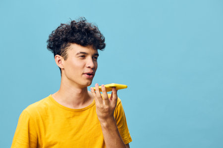 young man speaking into smartphone, cheerful expression, vibrant yellow shirt, bright blue background, modern lifestyle, technology concept, casual communication scenarioの写真素材