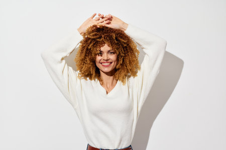 Happy young woman with curly hair wearing a cozy white sweater poses playfully with arms raised against a clean white background, exuding joy and confidenceの写真素材