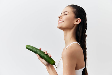 Woman holding a cucumber, smiling, with wet hair against a minimalistic white background, showcasing freshness and wellness concepts in a playful mannerの写真素材