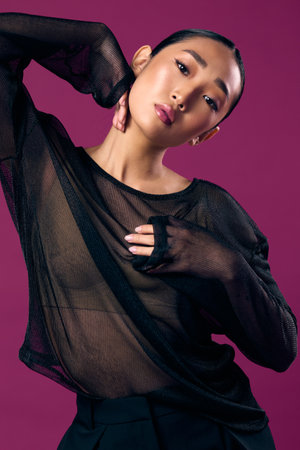 Fashion asian woman in a sheer top posing against a purple background with her hands on her hips and...