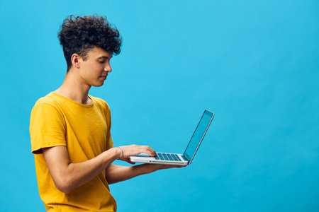 Young man using a laptop on a bright blue background, wearing a yellow t shirt, focusing on his work with a positive expression and modern technologyの写真素材