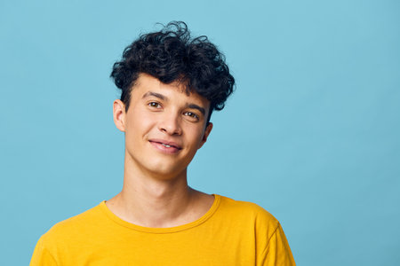 Young man with curly hair smiling against a light blue background, wearing a bright yellow shirt His cheerful expression evokes a sense of happiness and warmth in casual settingsの写真素材