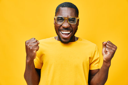 Excited young man celebrating success with a joyful expression, wearing stylish glasses and a yellow t shirt against a bright yellow background, full of vibrant energy and happinessの写真素材