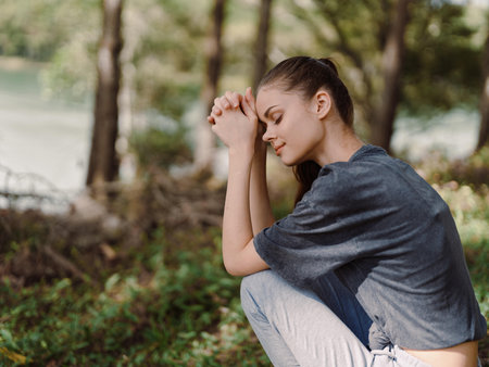 Thoughtful young woman sitting in the forest, hands clasped together, reflecting on life amidst...