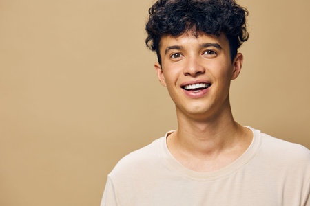 Happy young man smiling with curly hair in a light beige shirt against a warm beige background, representing joy and positivity in a casual settingの写真素材
