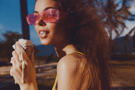A woman enjoying melting ice cream in a cone, wearing pink sunglasses, set against a sunny outdoor backdropの写真素材